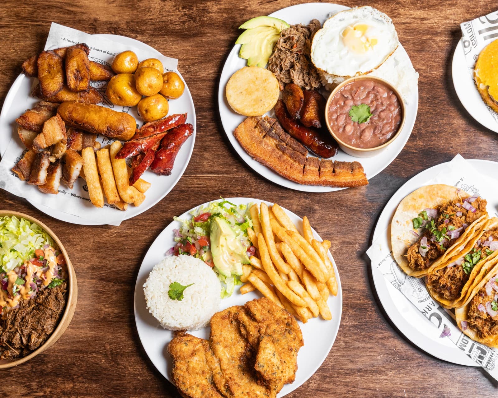 Two bowls of Lulada home-style plates — Colombian rice, beans, plantains, and pork belly on one side; a Mexican-style shredded beef bowl with pico de gallo on the other.