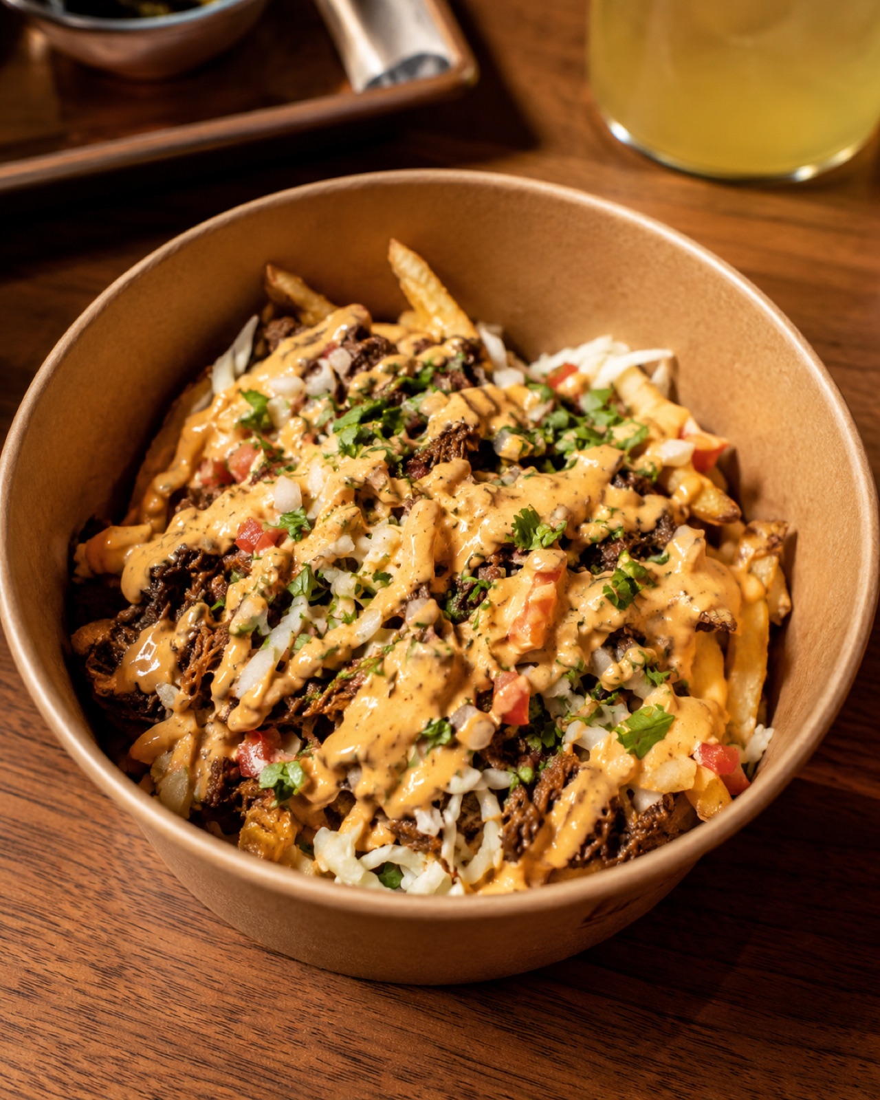 A bowl of birria fries on a café table, topped with shredded beef, cheese, and crema.